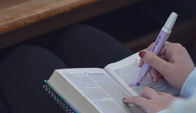 Two hands marking a line in an open book with a lilac highlighter.