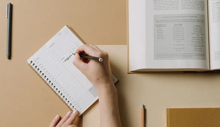 Hands writing in a planner on a desk, beside an open book