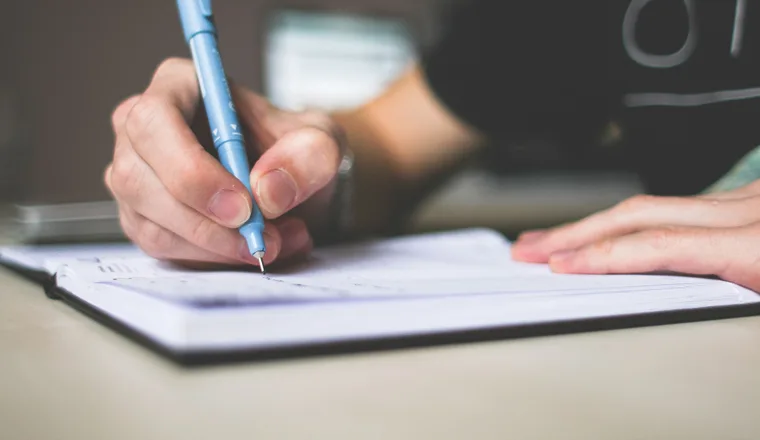 A hand writing with a blue pen in an open lined notebook on a desk.