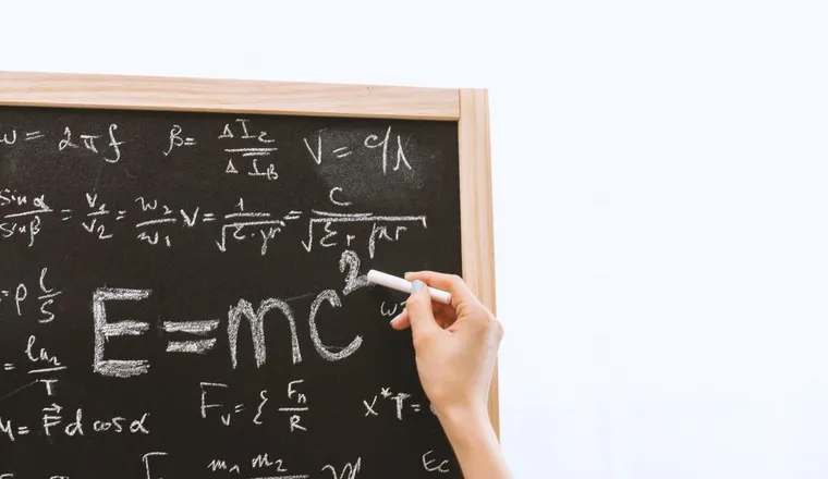 A blackboard covered in physics equations, a hand writing among them