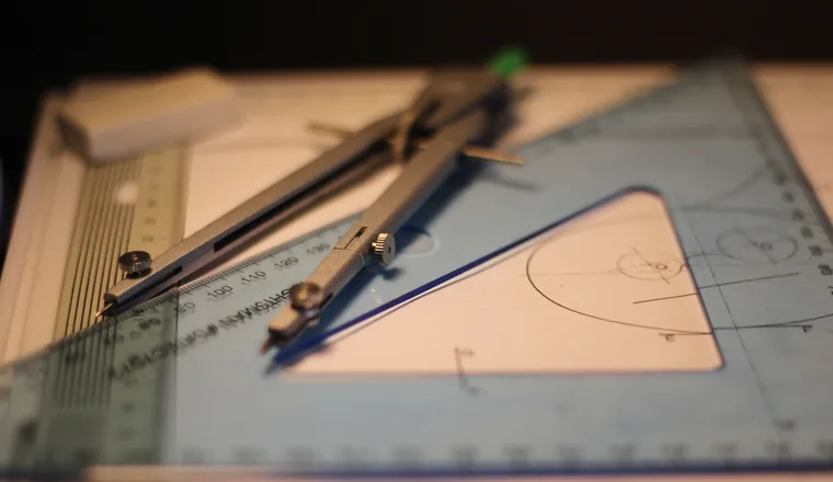A metal compass and a clear blue set square resting on a sheet of drawing paper.