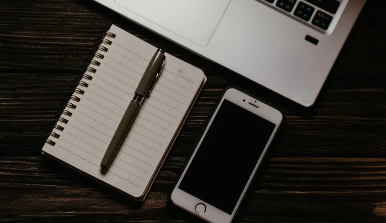 A laptop, a phone and a spiral notebook laid out on a dark wooden table