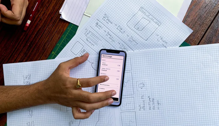 A hand and a phone resting over sheets of graph paper on a wooden desk