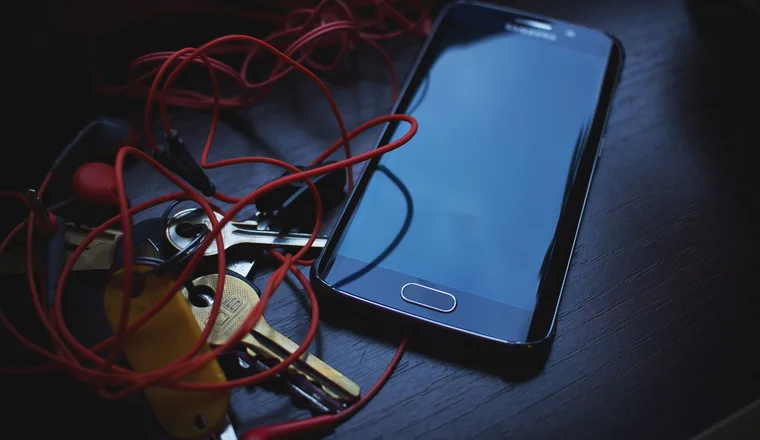 A phone and a set of keys on a dark table, earphones coiled beside them
