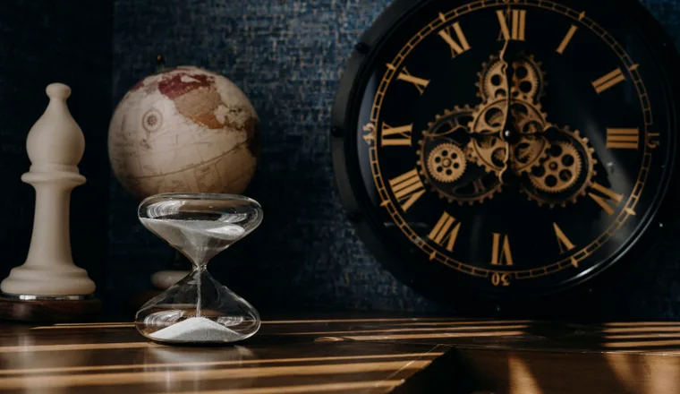 An hourglass and an ornate clock on a dark table